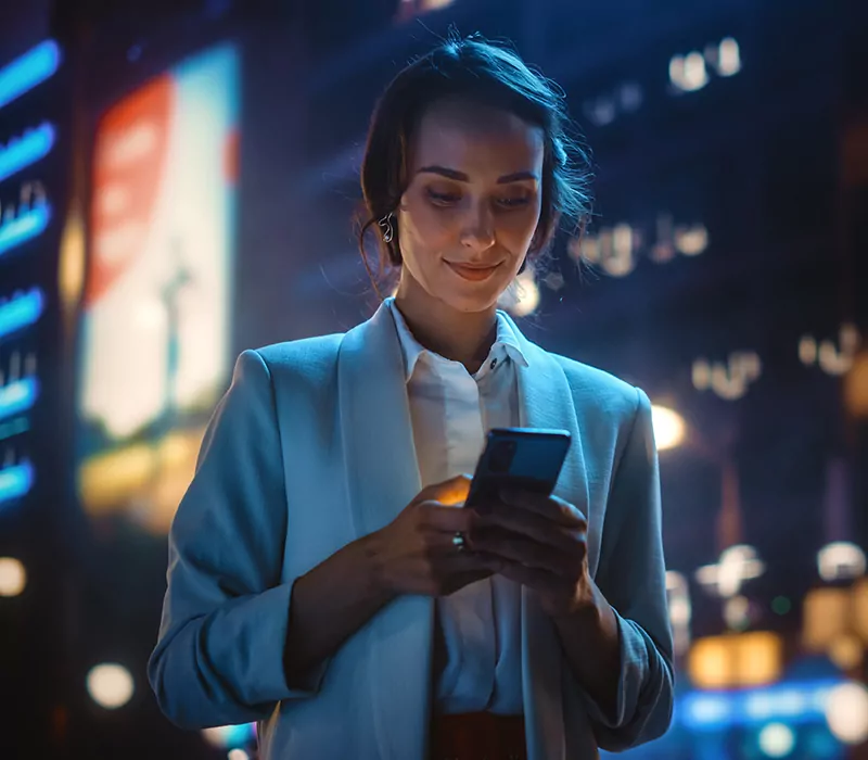 beautiful young woman using smartphone walking through night city street full of neon light. portrait of gorgeous smiling female using mobile phone.