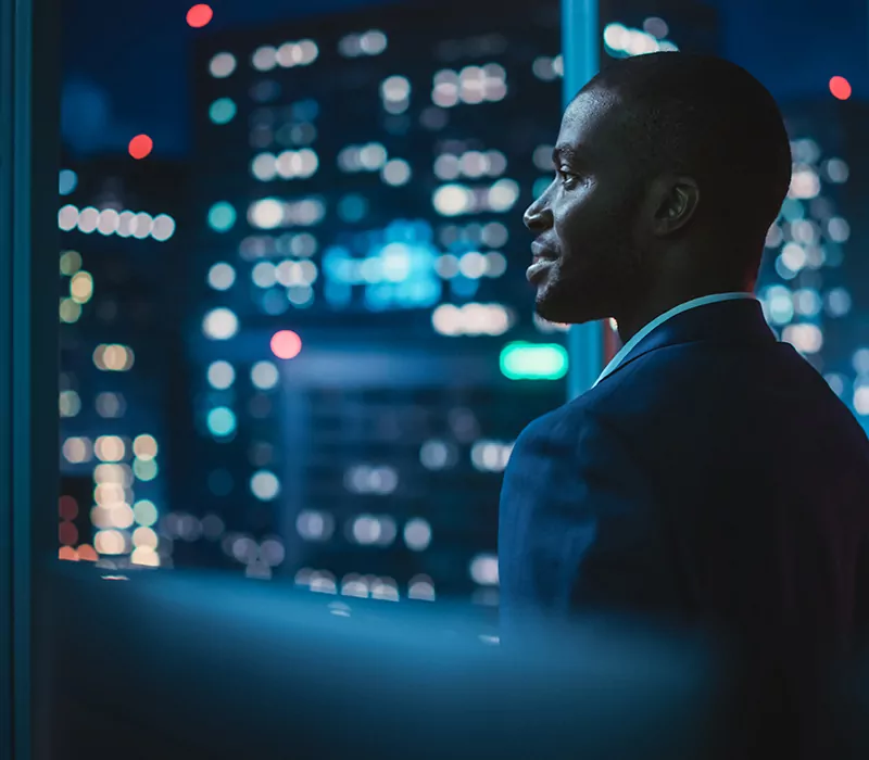 successful businessman looking out of the window on late evening. modern hedge fund investor enjoying successful life. urban view with down town street with skyscrapers at night with neon lights.